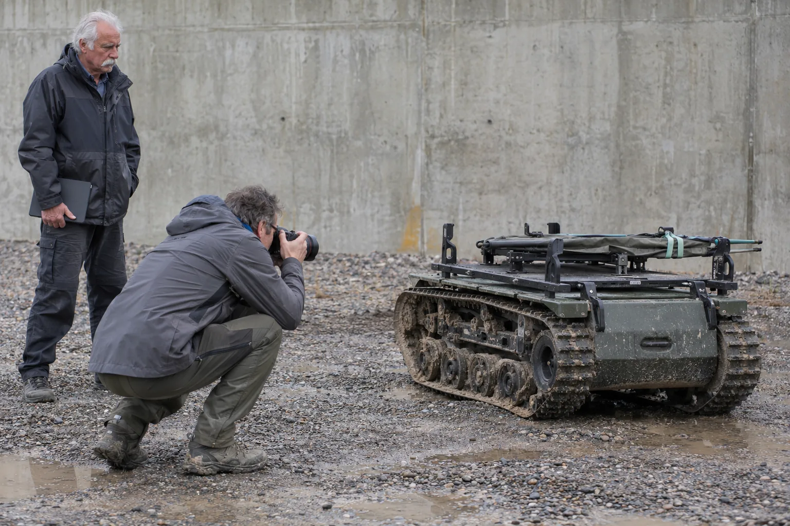 Photographer shooting a tracked ground robot carrying a stretcher frame on a bare gravel test pad